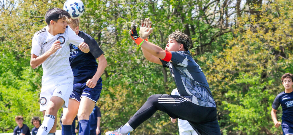 Youth soccer players contest a header while a goalkeeper jumps to block the ball during an outdoor match.